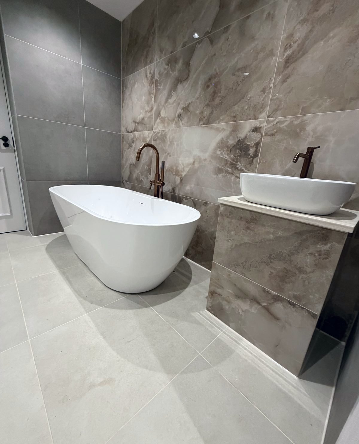 The same room finished: freestanding bath and vessel basin vanity in marble-effect tiles with copper taps